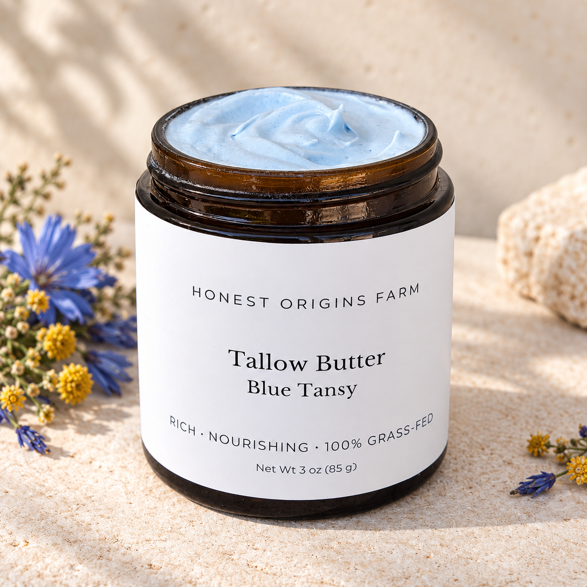 A jar of Honest Origins Farm Tallow Butter in Blue Tansy scent, filled with light blue cream and set in a sandy, natural setting with small wildflowers.