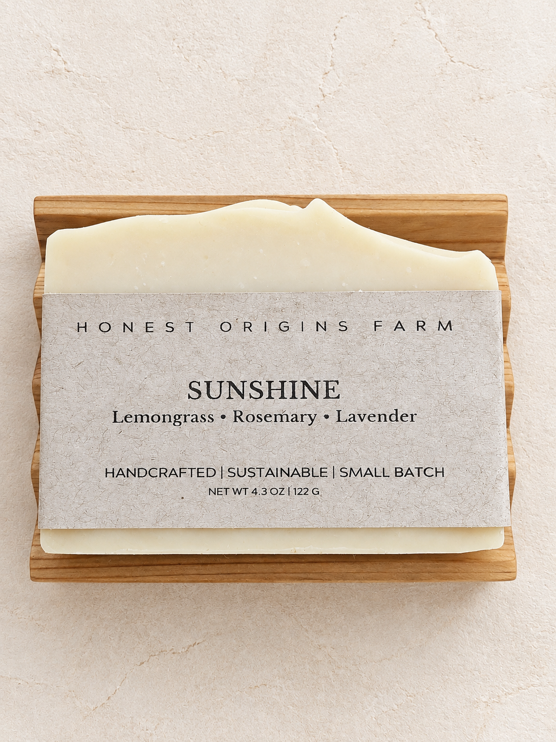 A bar of Honest Origins Farm 'Sunshine' handmade soap in a light wooden soap dish, set against a textured beige background. The soap bar is wrapped in a neutral gray paper sleeve showing the scent notes of Lemongrass, Rosemary, and Lavender.