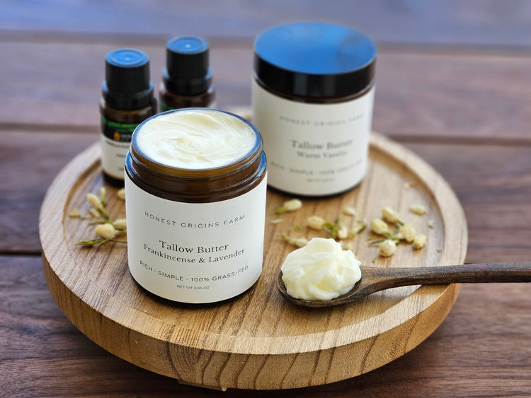 Tallow body butter on a wooden tray with small jars of essential oils, a spoon with body butter, and jasmine flowers