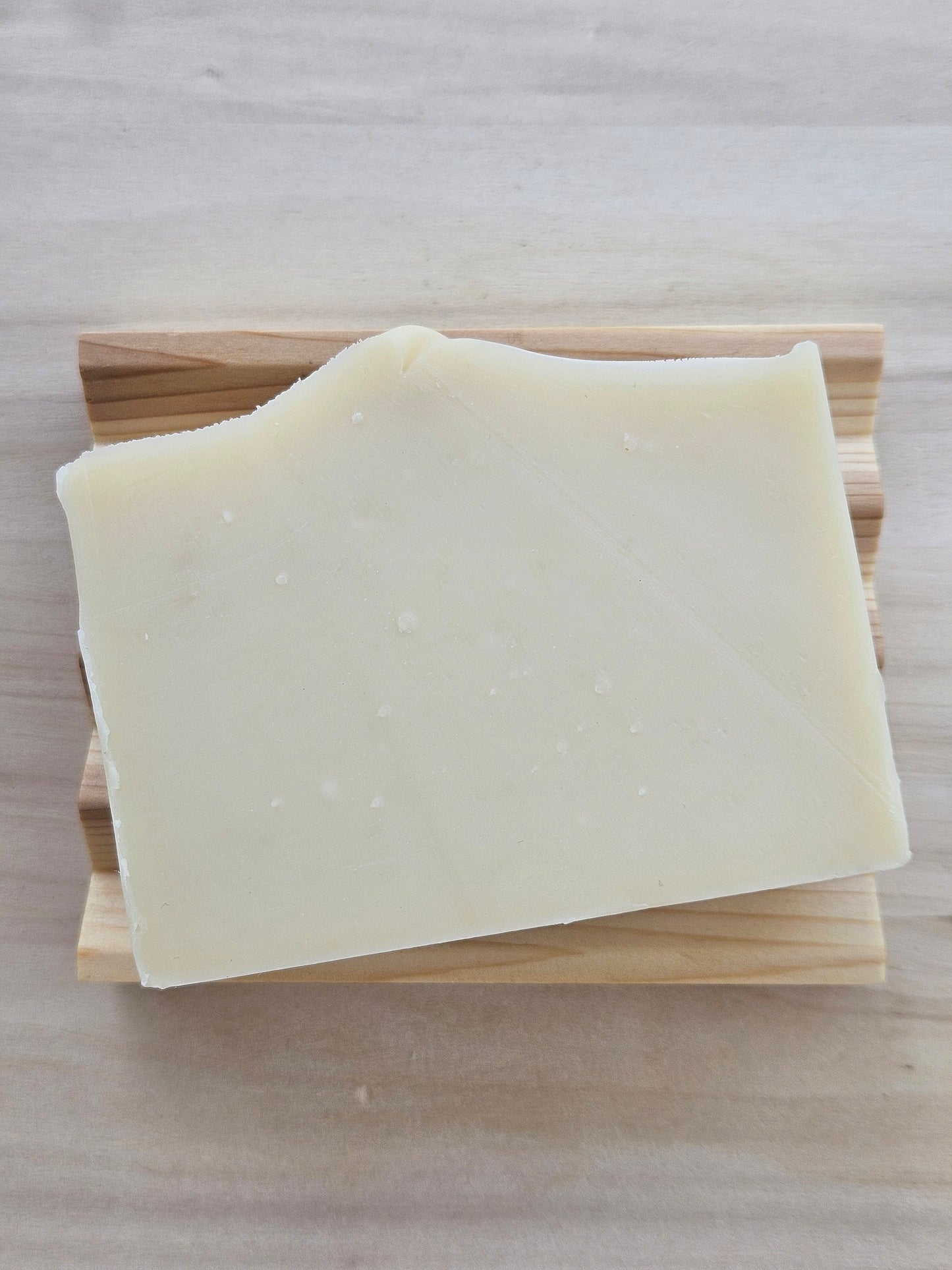 A thick, solid bar of pale cream-colored soap resting on a cedar soap dish, set against a light wood grain background.
