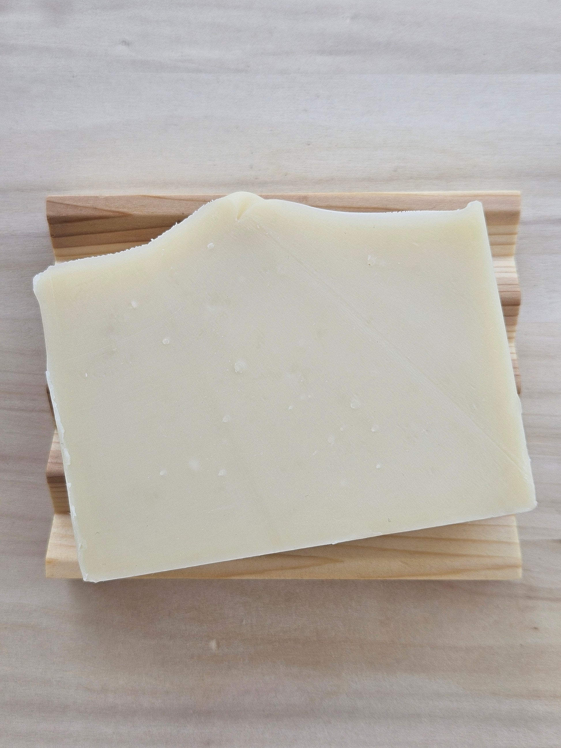 A thick, solid bar of pale cream-colored soap resting on a cedar soap dish, set against a light wood grain background.