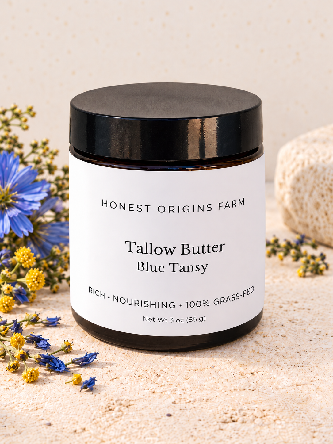 A jar of Honest Origins Farm Tallow Butter Blue Tansy, 3 oz, with a black lid and white label, styled on a sandy surface with blue and yellow wildflowers.