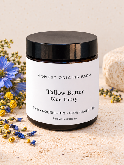A jar of Honest Origins Farm Tallow Butter Blue Tansy, 3 oz, with a black lid and white label, styled on a sandy surface with blue and yellow wildflowers.