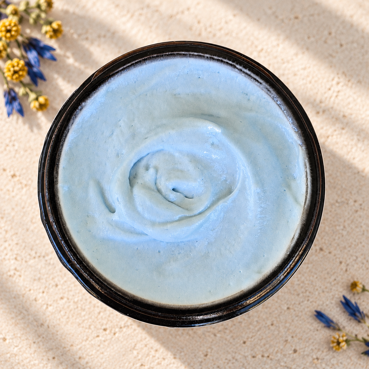 A top-down view of a amber cosmetic jar filled with light blue cream, placed on a sandy beige surface with small dried flowers around it.