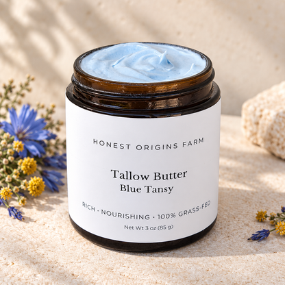 A jar of Honest Origins Farm Tallow Butter in Blue Tansy scent, filled with light blue cream and set in a sandy, natural setting with small wildflowers.