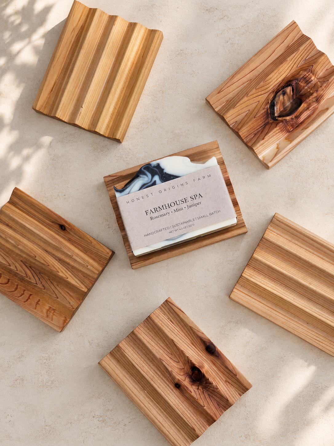 Five rectangular cedar soap dishes with natural grain patterns are arranged on a light beige textured surface with soft leaf shadows. A small card reading 'FARMHOUSE SPA' is placed on one of the dishes.