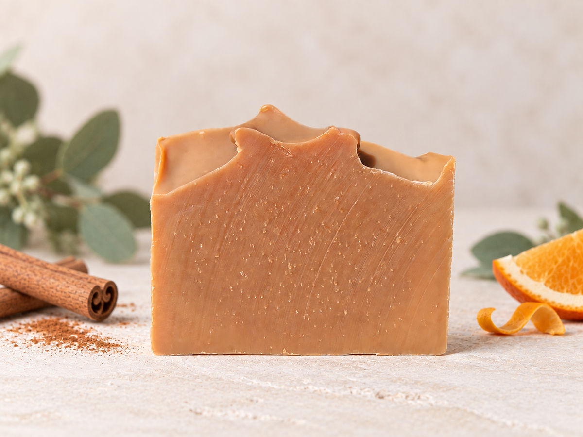 A rectangular bar of warm tan soap with a rustic, hand-drawn top edge, styled on a sandy surface with cinnamon sticks, orange peel, and small flowers in the background.