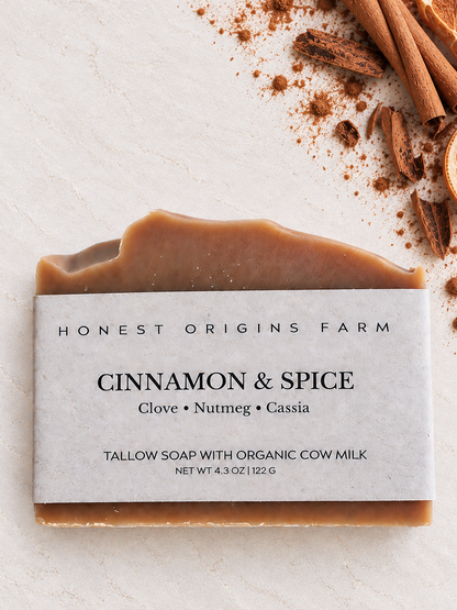 A bar of Honest Origins Farm Cinnamon & Spice tallow soap with organic cow milk, wrapped in a white paper label. The soap has a warm brown color and is styled on a light textured background with cinnamon sticks and cinnamon.