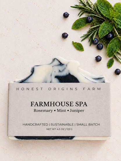 A bar of handmade soap from Honest Origins Farm, labeled 'Farmhouse Spa', styled on a textured cream background with rosemary, mint, and blueberries.