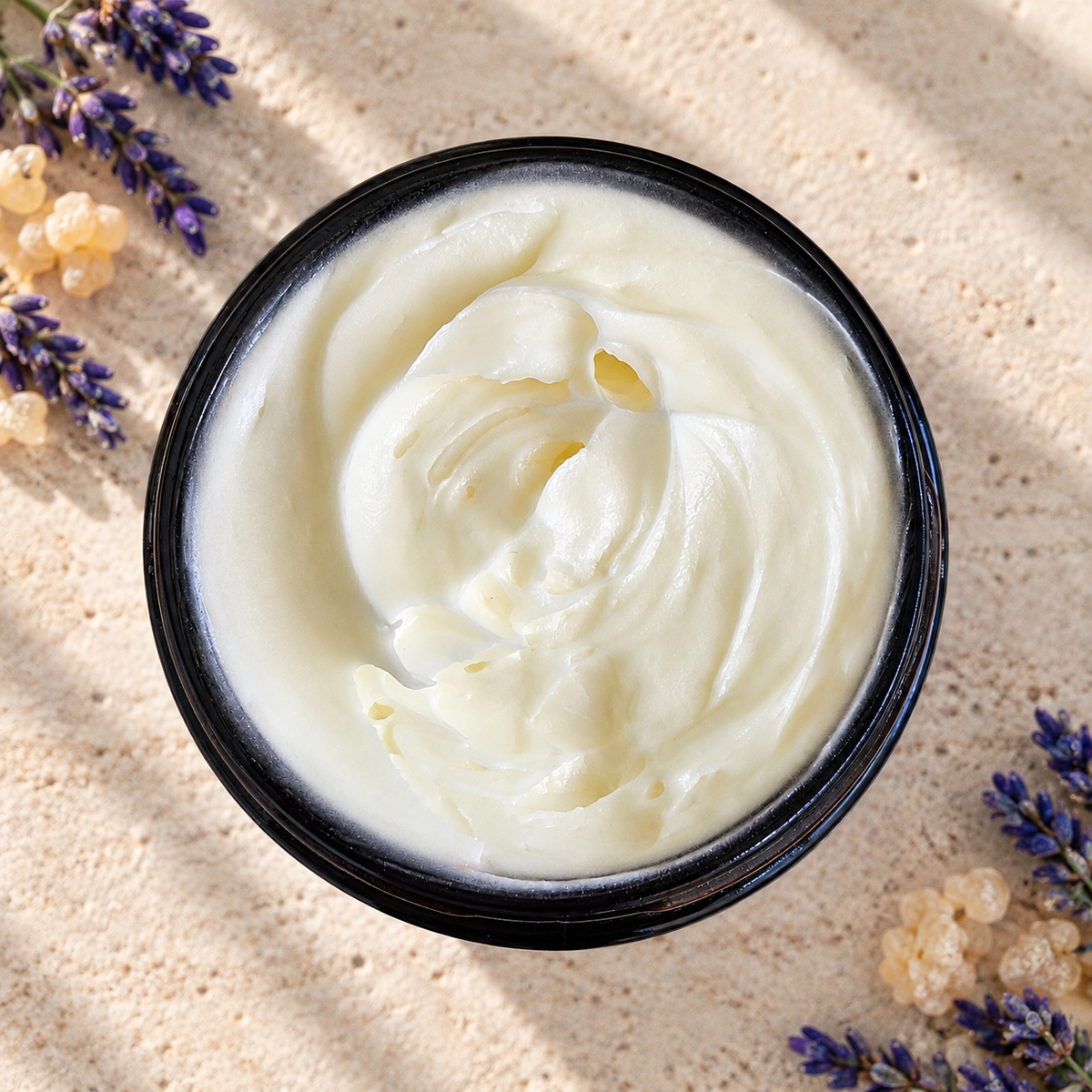 A top-down view of a black jar filled with lotion, surrounded by lavender sprigs and small beige botanicals on a textured sandy background.