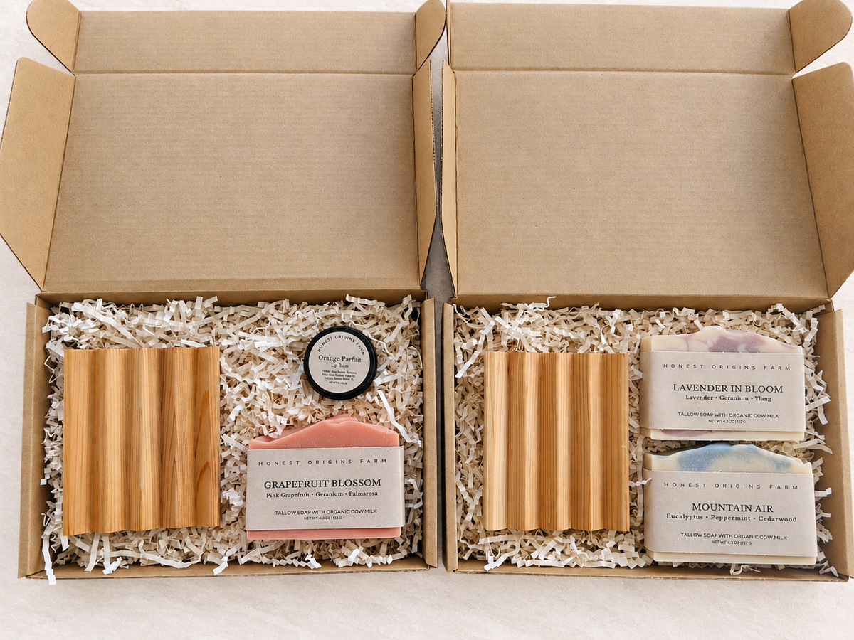 Two open kraft gift boxes filled with white paper shred, each containing a cedar soap dish, a small round tin, and two rectangular soaps labeled Grapefruit Blossom and Lavender in Bloom, with Mountain Air also visible.