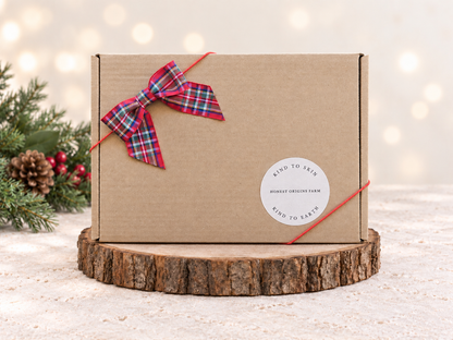 A square kraft cardboard gift box with a red and blue plaid bow and red string, featuring a round white sticker that reads 'KIND TO SKIN / MONEY DESIGNS FARM / KIND TO EARTH', displayed on a rustic wood slice with holiday greenery and warm bokeh lights in the background.