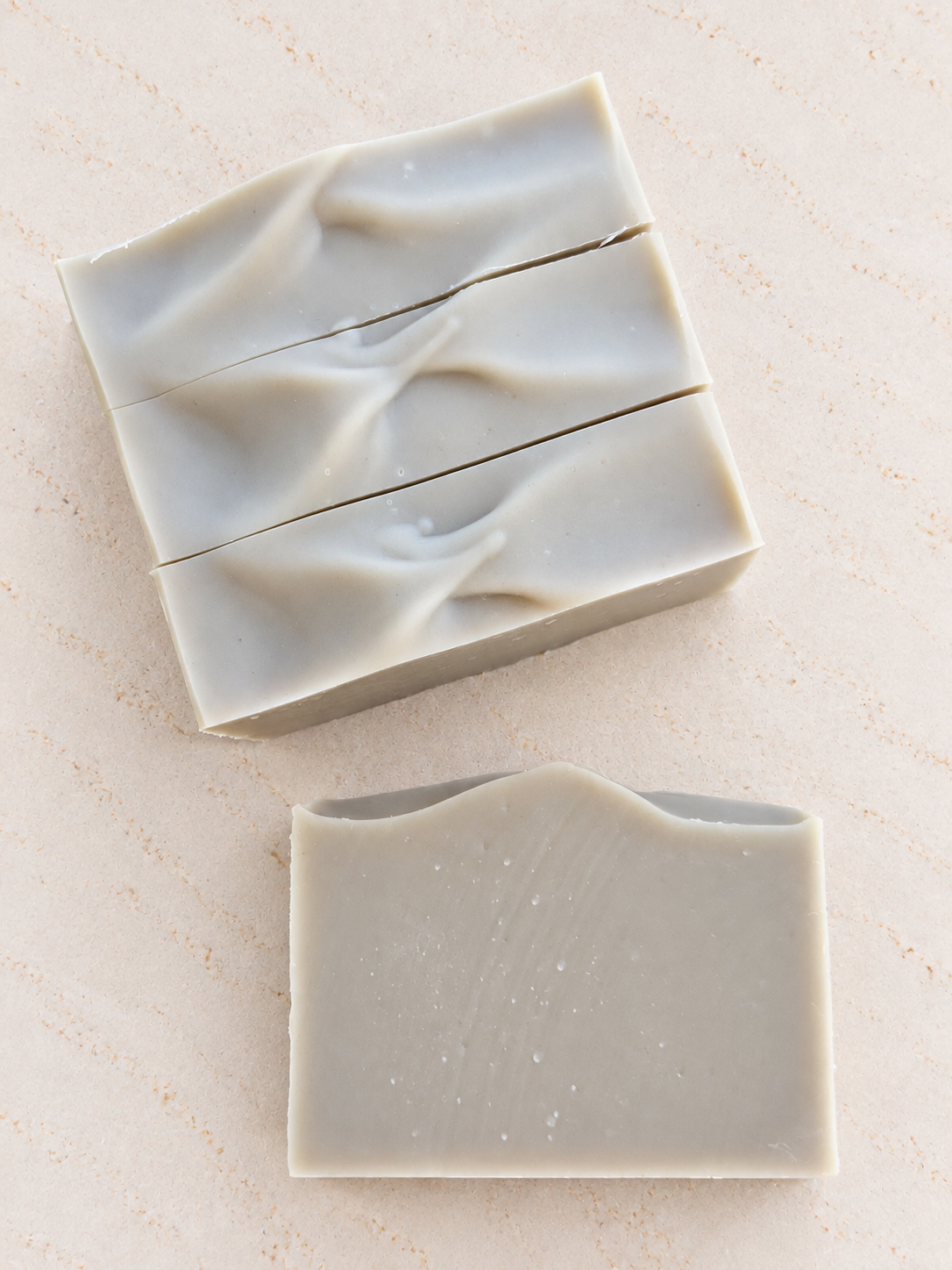 Three rectangular bars of handmade soap in a light green color, arranged on a textured beige surface, with one bar cut horizontally in the middle.