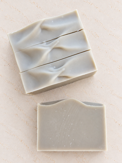 Three rectangular bars of handmade soap in a light green color, arranged on a textured beige surface, with one bar cut horizontally in the middle.