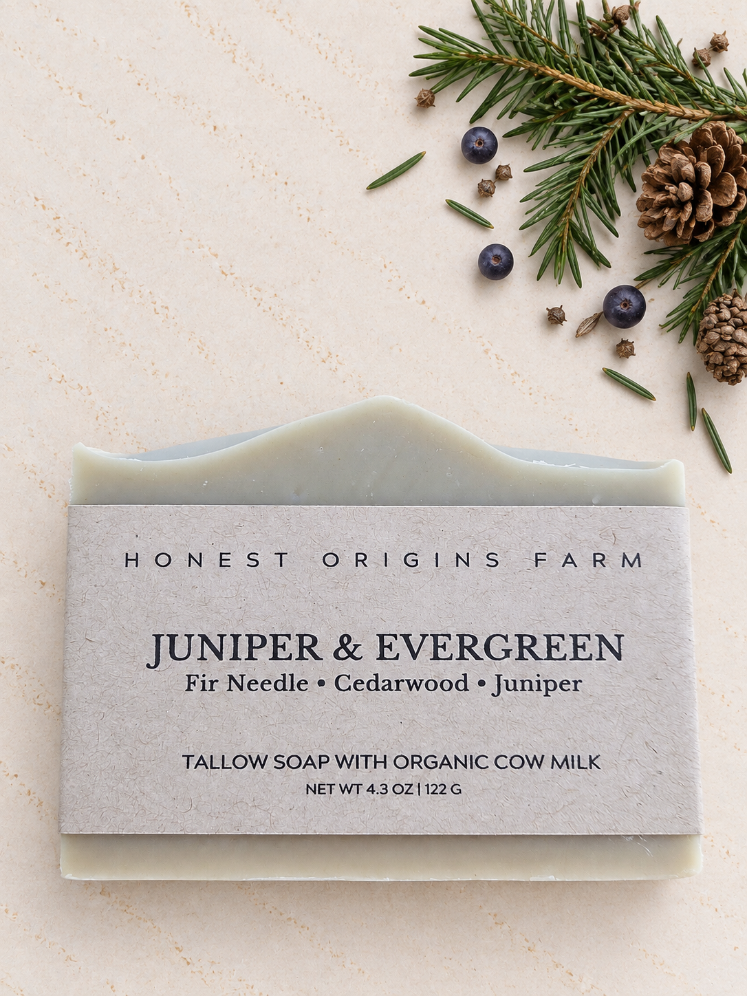 A bar of Honest Origins Farm Juniper & Evergreen tallow soap with an organic cow milk scent, displayed on a textured beige background with flat-lay holiday props including pine needles, pinecones, and juniper berries.