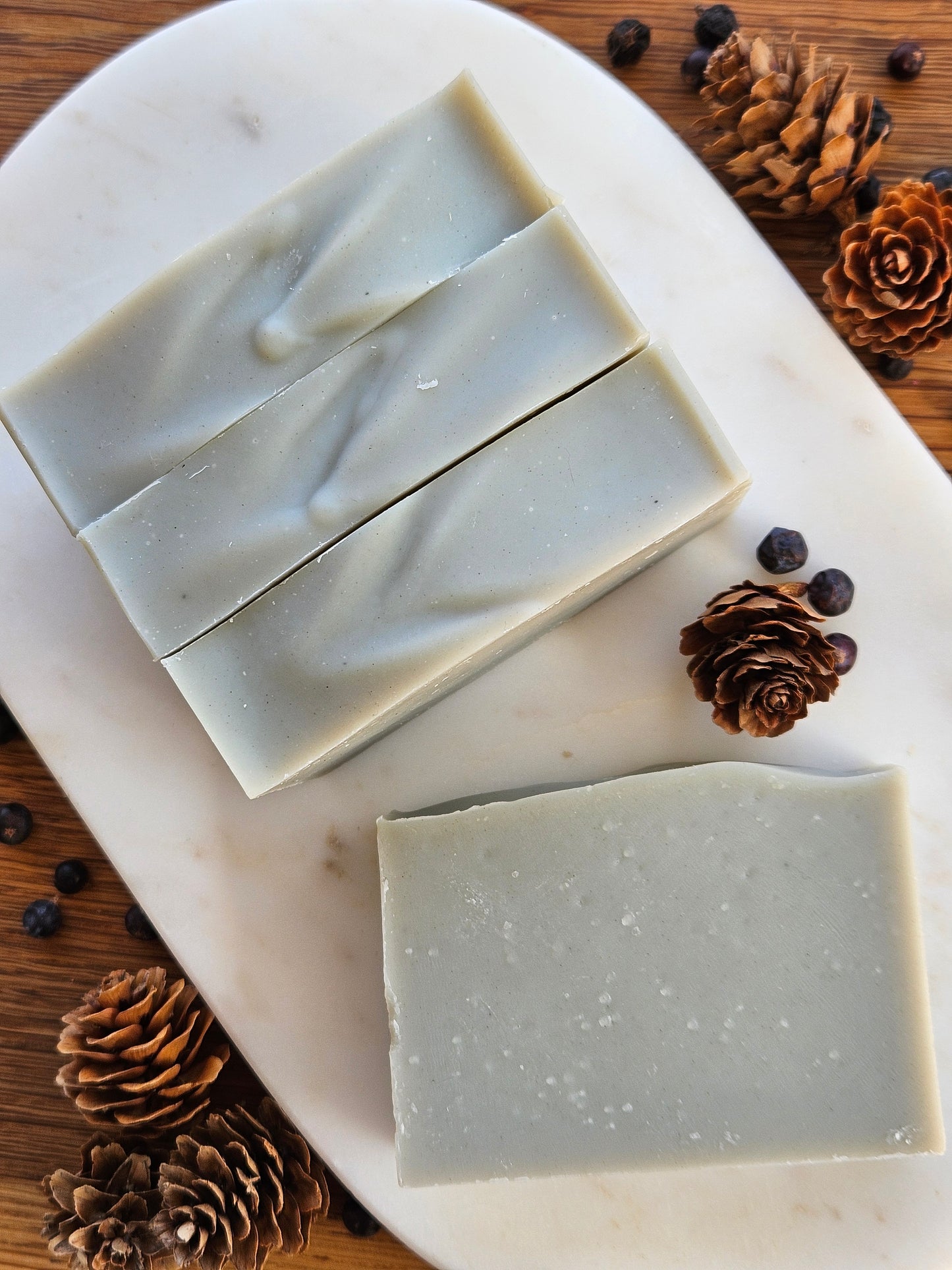 Three bars of soap on a marble slab with pine cones and berries on a wooden surface
