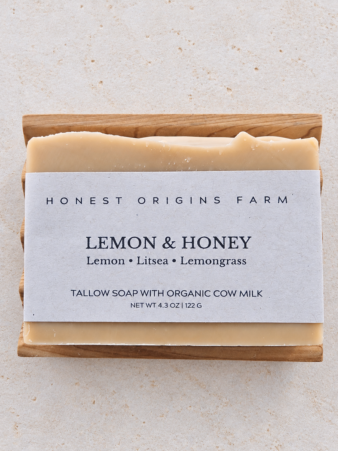 A rectangular bar of Honest Origins Farm Lemon & Honey tallow soap resting on a light wood block, with a white paper label, on a textured beige stone background.
