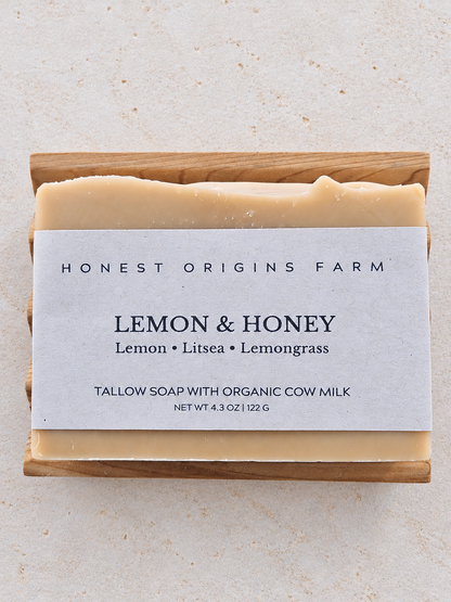 A rectangular bar of Honest Origins Farm Lemon & Honey tallow soap resting on a light wood block, with a white paper label, on a textured beige stone background.