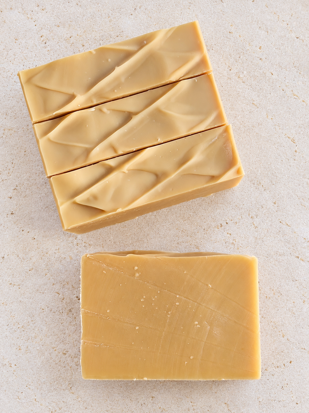 Three rectangular bars of pale yellow soap with textured, swirled tops, arranged on a light beige stone-like surface. One bar lays flat to show the soap texture once cut.
