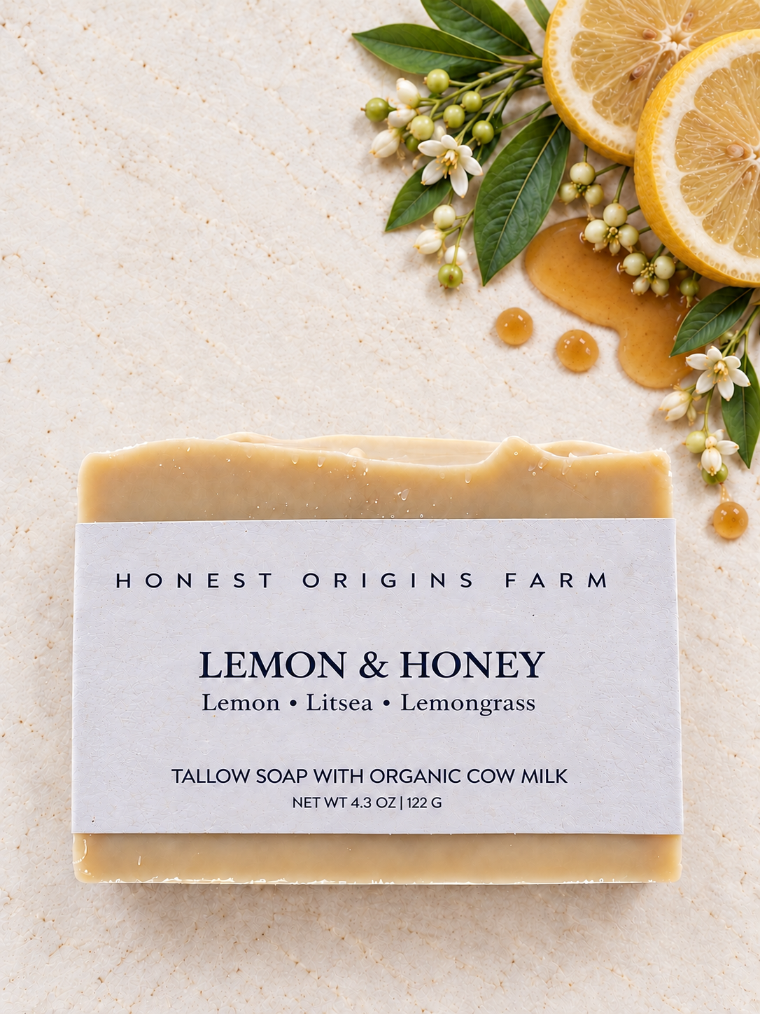 A rectangular bar of Honest Origins Farm Lemon & Honey tallow soap with a white paper label, styled on a light beige textured surface with lemon slices, honey, green leaves, and small white flowers.