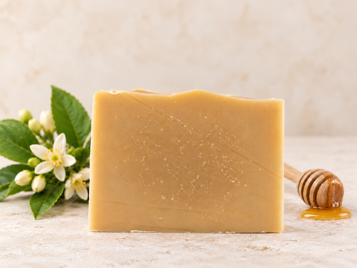 A rectangular bar of handmade honey-colored soap stands on a light stone surface, flanked by white flowers with green leaves on the left and a wooden honey dipper with honey on the right.