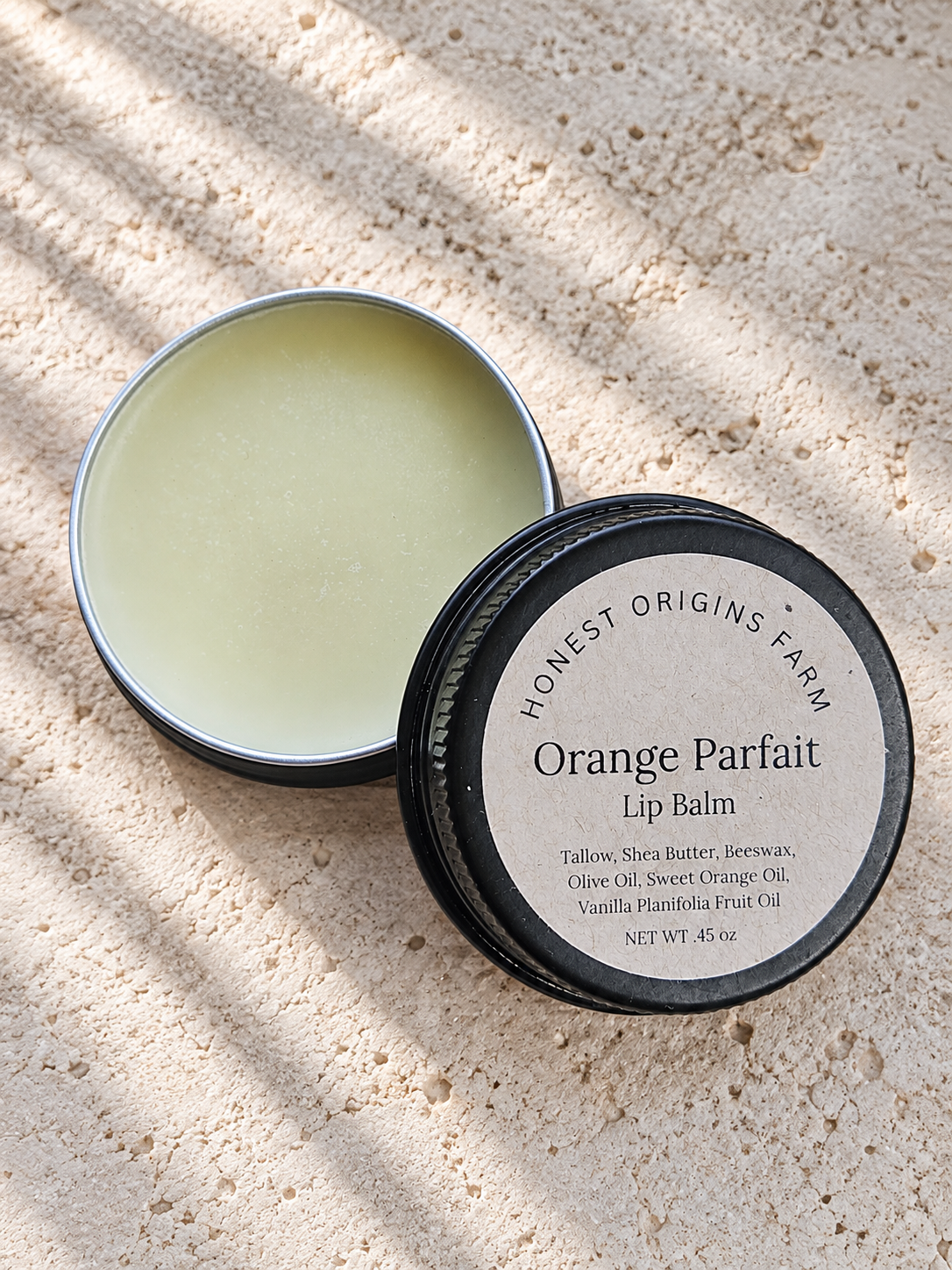 An open round metal tin of Honest Origins Farm Orange Parfait Lip Balm sits on a sandy surface with dappled sunlight and shadows. The lid, featuring a beige label with black text, rests beside the tin.