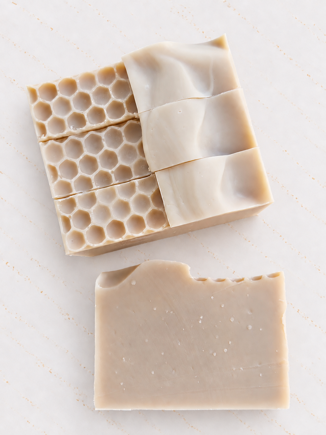 Three bars of handmade soap in light beige tones, including one bar with a honeycomb pattern, arranged on a light marble background.