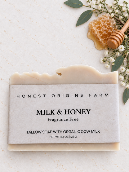 A bar of Honest Origins Farm Milk & Honey fragrance-free tallow soap with organic cow milk, styled on a light marble surface with a honeycomb, wooden honey dipper, and small white flowers.