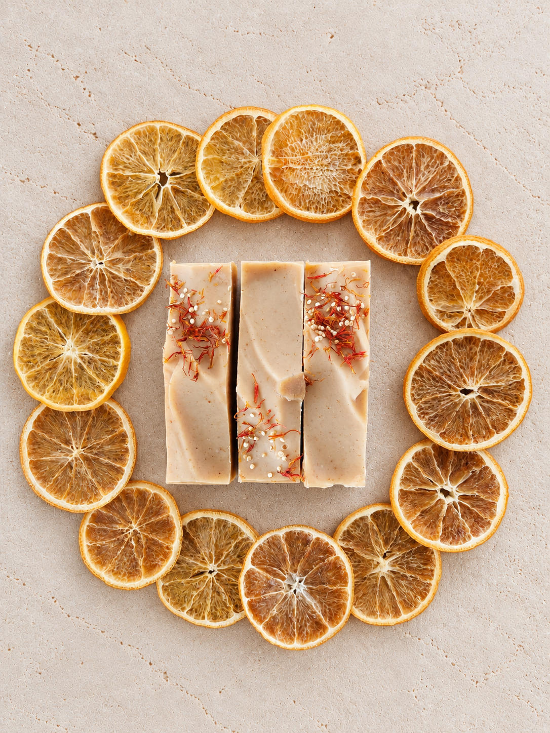 Three rectangular bars of orange-cream colored soap with dried orange botanical inclusions, arranged in the center of a circular arrangement of dried orange slices on a light beige textured surface.