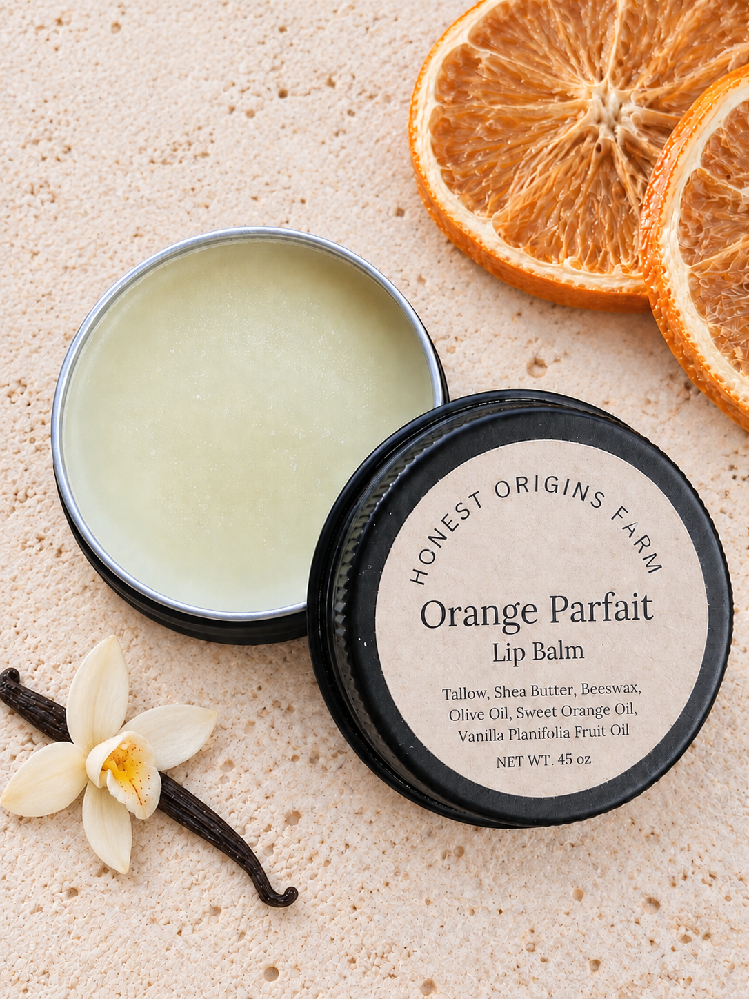 An open round tin of Honest Origins Farm Orange Parfait Lip Balm sits beside its black lid on a light sandy surface, with dried orange slices and a vanilla flower nearby.