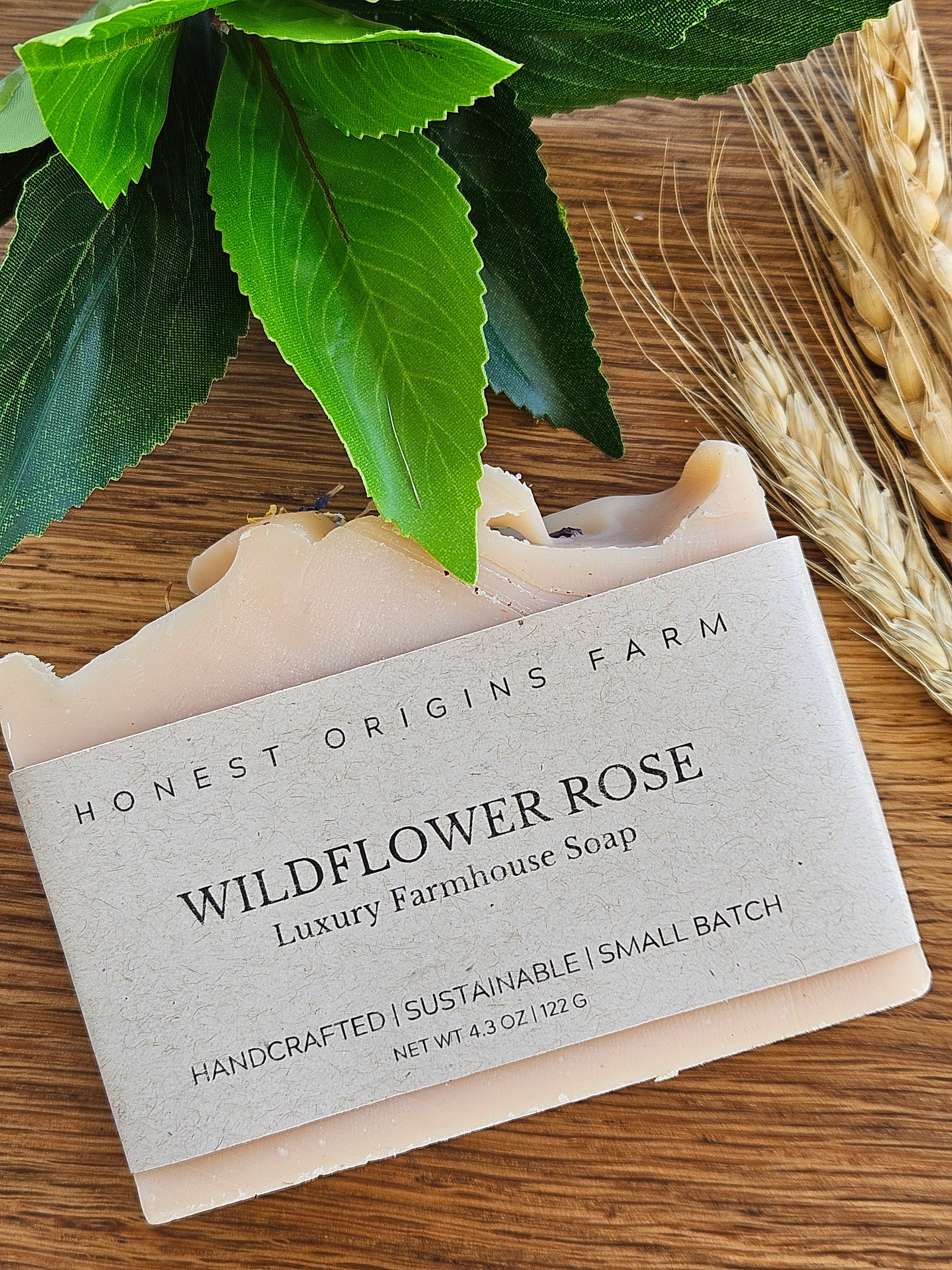 Bar of soap with a label on a wooden surface, surrounded by green leaves and wheat stalks.