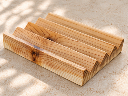 A cedar soap dish with visible grain, shown outdoors on sandy ground with dappled sunlight and shadows.