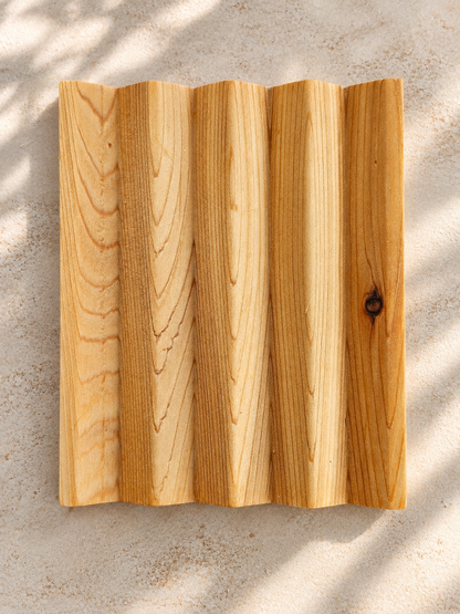 A cedar soap dish with a wavy, corrugated edge and visible wood grain, photographed from above on a light textured surface with soft leaf shadows.