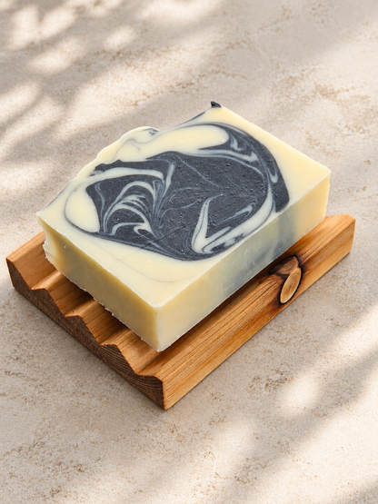 A rectangular bar of handmade soap with a cream and black marbled swirl pattern, resting on a cedar soap dish against a textured beige background with soft leaf shadows.