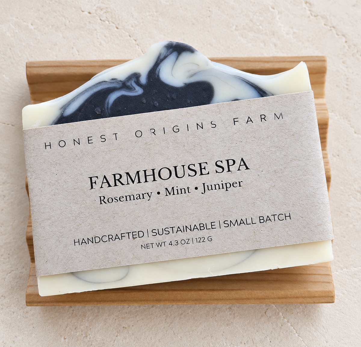 A handcrafted bar of soap from Honest Origins Farm, labeled 'Farmhouse Spa' with rosemary, mint, and juniper scents. The soap has a cream base with a black and cream swirled top layer, resting on a soap dish against a textured beige background.