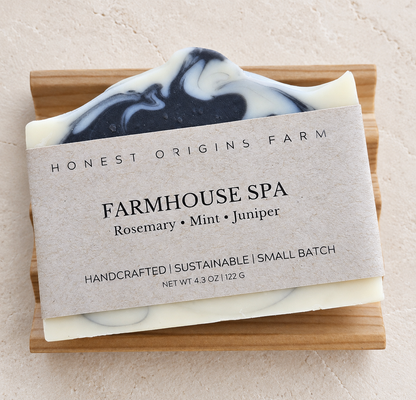 A handcrafted bar of soap from Honest Origins Farm, labeled 'Farmhouse Spa' with rosemary, mint, and juniper scents. The soap has a cream base with a black and cream swirled top layer, resting on a soap dish against a textured beige background.