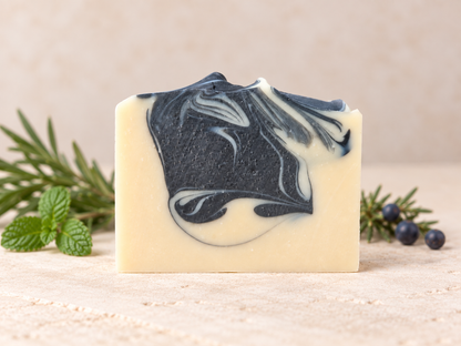 A rectangular bar of handmade soap with a cream-colored base and a black  marbled swirl pattern, styled on a neutral beige surface with rosemary and blueberries.