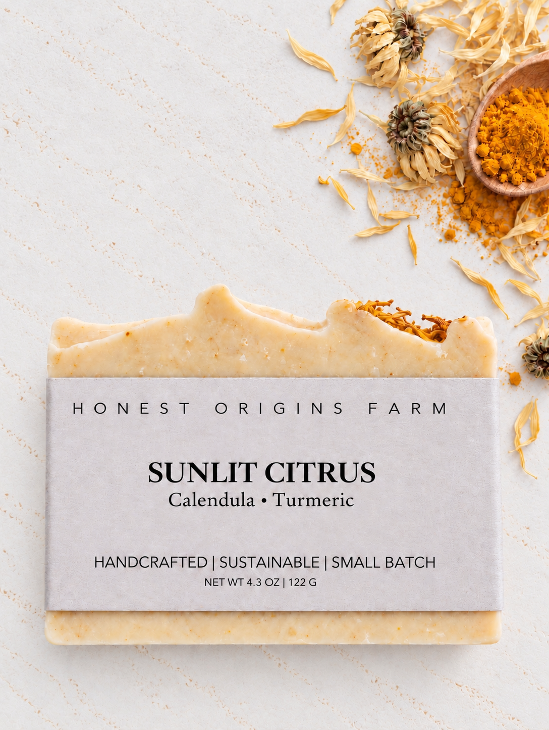 A rectangular bar of Honest Origins Farm Sunlit Citrus soap with a light gray paper label, styled on a textured off-white surface with dried calendula flowers, a small wooden spoon of orange-yellow powder, and scattered dried petals.