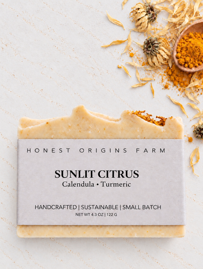 A rectangular bar of Honest Origins Farm Sunlit Citrus soap with a light gray paper label, styled on a textured off-white surface with dried calendula flowers, a small wooden spoon of orange-yellow powder, and scattered dried petals.