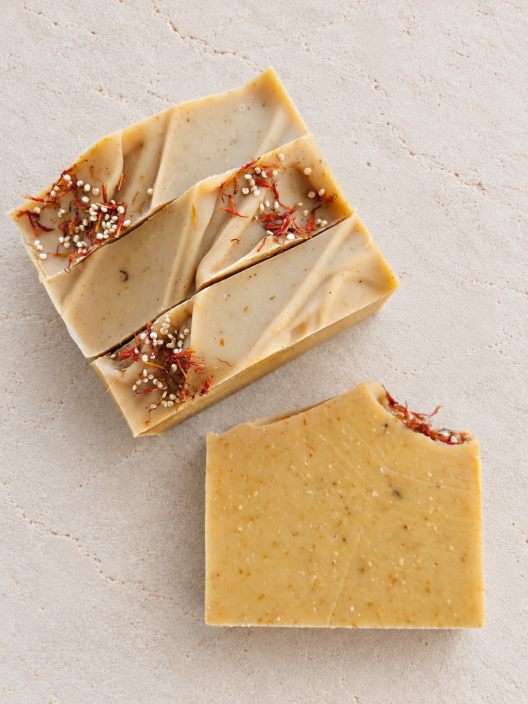 Three rectangular bars of handmade soap in warm beige and golden yellow tones, decorated with dried red flowers and white seeds, arranged on a textured light beige surface.