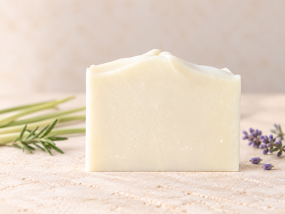A rectangular bar of creamy off-white soap standing upright on a light textured surface, with green herb sprigs on the left and small purple lavender flowers on the right, against a soft neutral background.
