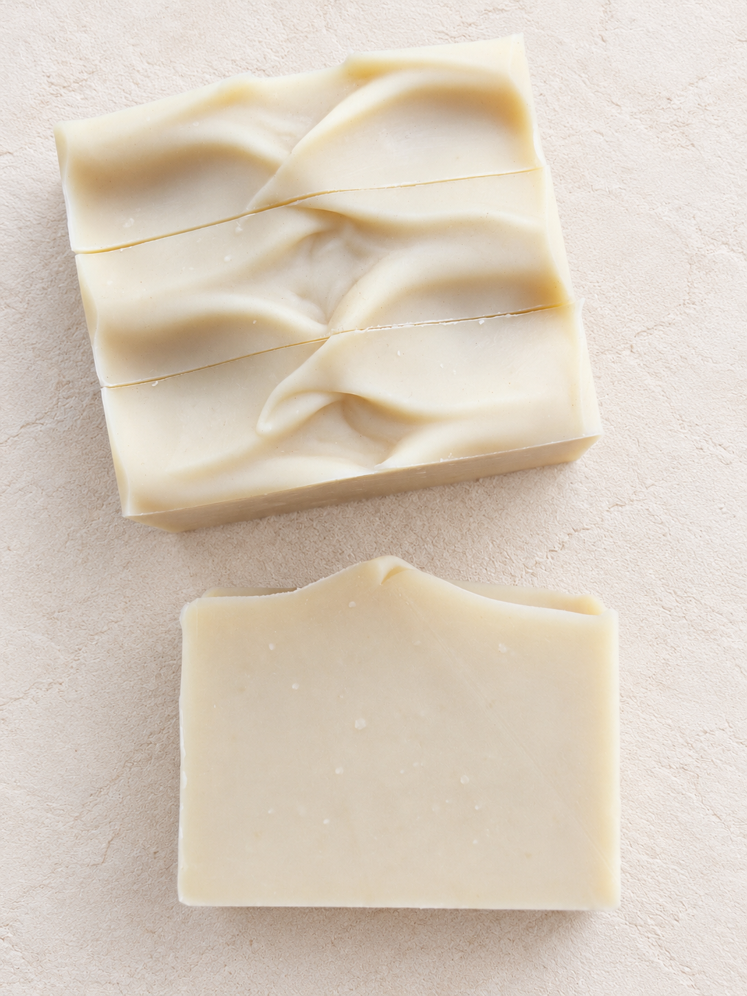 Three rectangular bars of handmade soap in a warm cream color, displayed on a textured light beige surface. One bar lying on its side in front showing the side of the soap.