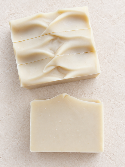 Three rectangular bars of handmade soap in a warm cream color, displayed on a textured light beige surface. One bar lying on its side in front showing the side of the soap.