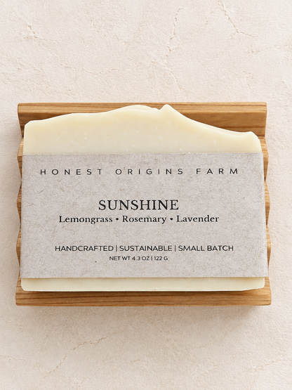 A bar of Honest Origins Farm 'Sunshine' handmade soap in a light wooden soap dish, set against a textured beige background. The soap bar is wrapped in a neutral gray paper sleeve showing the scent notes of Lemongrass, Rosemary, and Lavender.