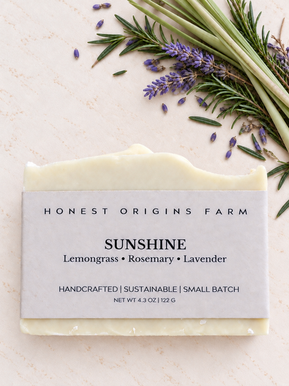 A handcrafted bar of soap from Honest Origins Farm, labeled 'Sunshine' with notes of lemongrass, rosemary, and lavender, styled on a light textured background with fresh rosemary and lavender sprigs.
