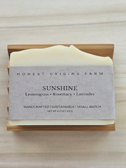 A bar of Honest Origins Farm 'Sunshine' handmade soap in a light wood soap dish, featuring a textured gray paper label with the scent notes 'Lemongrass • Rosemary • Lavender'.