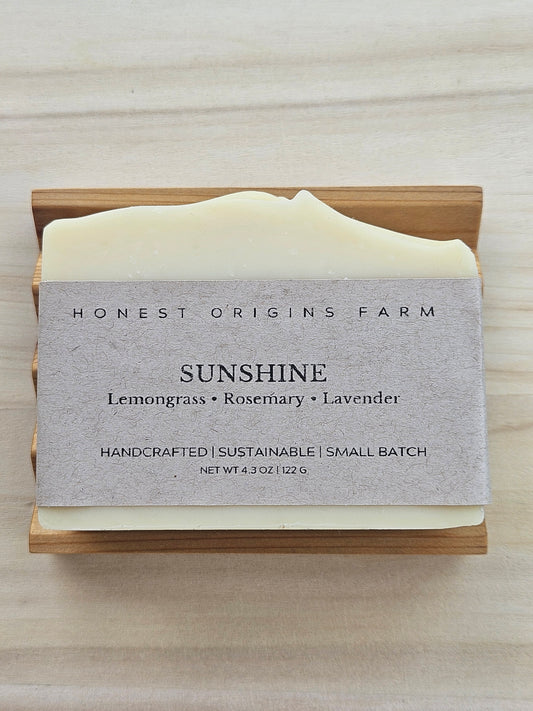 A bar of Honest Origins Farm 'Sunshine' handmade soap in a light wood soap dish, featuring a textured gray paper label with the scent notes 'Lemongrass • Rosemary • Lavender'.