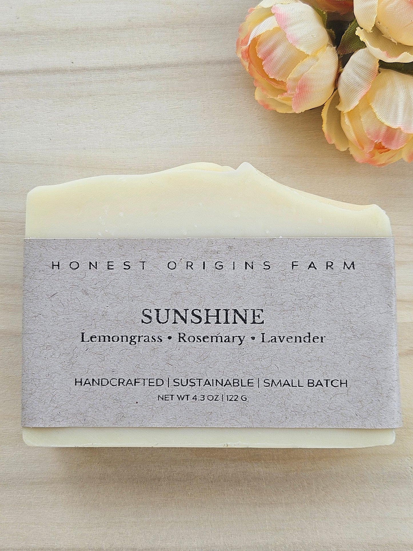 A bar of Honest Origins Farm 'Sunshine' handmade soap with a light gray paper label, displayed on a light wood background with peach roses in the top right corner.