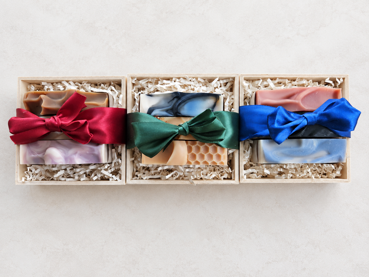 Three wooden gift boxes filled with artisanal handmade soaps, arranged side by side on a light beige surface. Each box is wrapped with a large satin ribbon in red, green, and blue, respectively.
