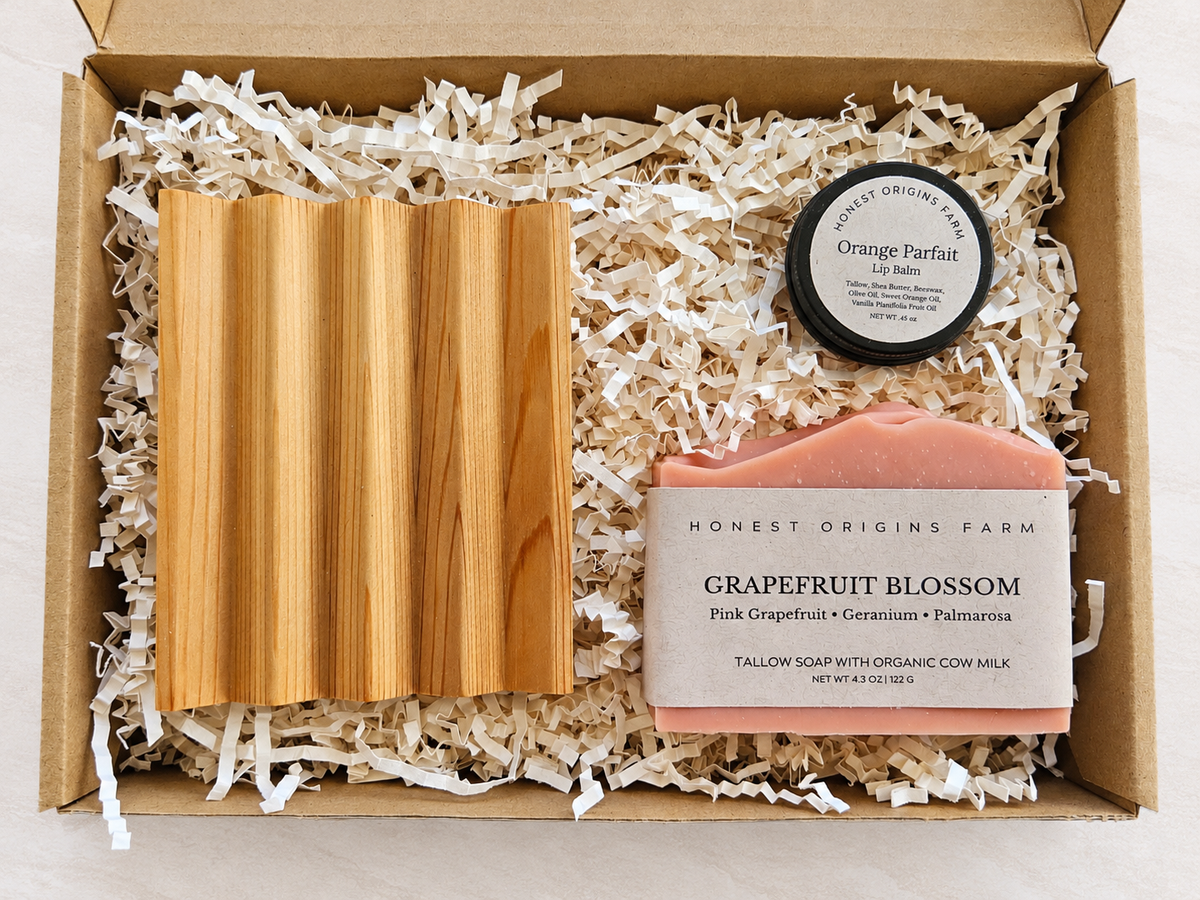 An open cardboard gift box filled with shredded paper packaging, containing a pink grapefruit blossom soap bar, a cedar soap dish and a small round tin labeled Orange Parfait Lip Balm by Honest Origins Farm.